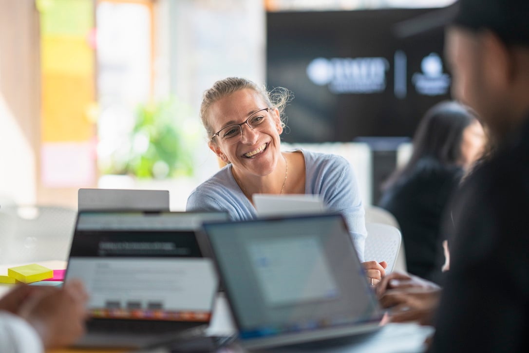 Picture of Steph smiling. 3 MacBook in the foreground.
Steph is wearing glasses and that same light blue top.